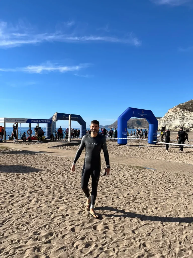 Pablo con el neopreno puesto caminando por la arena de la playa de Agua Amarga, sonriendo, con los arcos de la salida y el mar detrás.