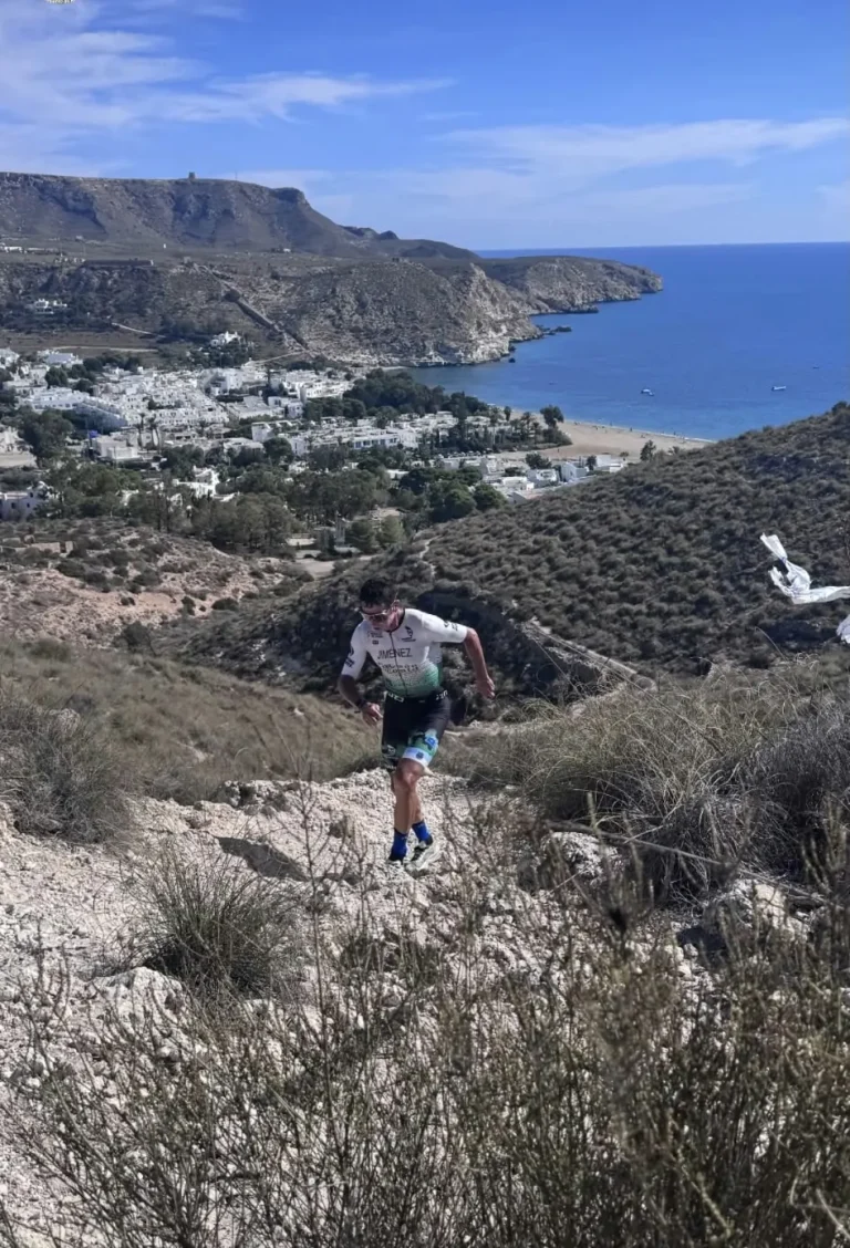 Un triatleta sube un sendero de piedra suelta entre matorrales en la carrera a pie, con Agua Amarga y el mar al fondo.